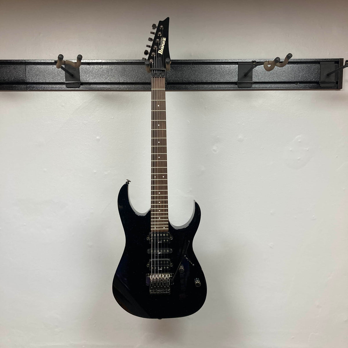 An Ibanez Prestige RG1570 Cosmic Blue electric guitar with case sits on a wall-mounted rack against a plain white wall, highlighting its vivid blue finish and signature Wizard Prestige neck.