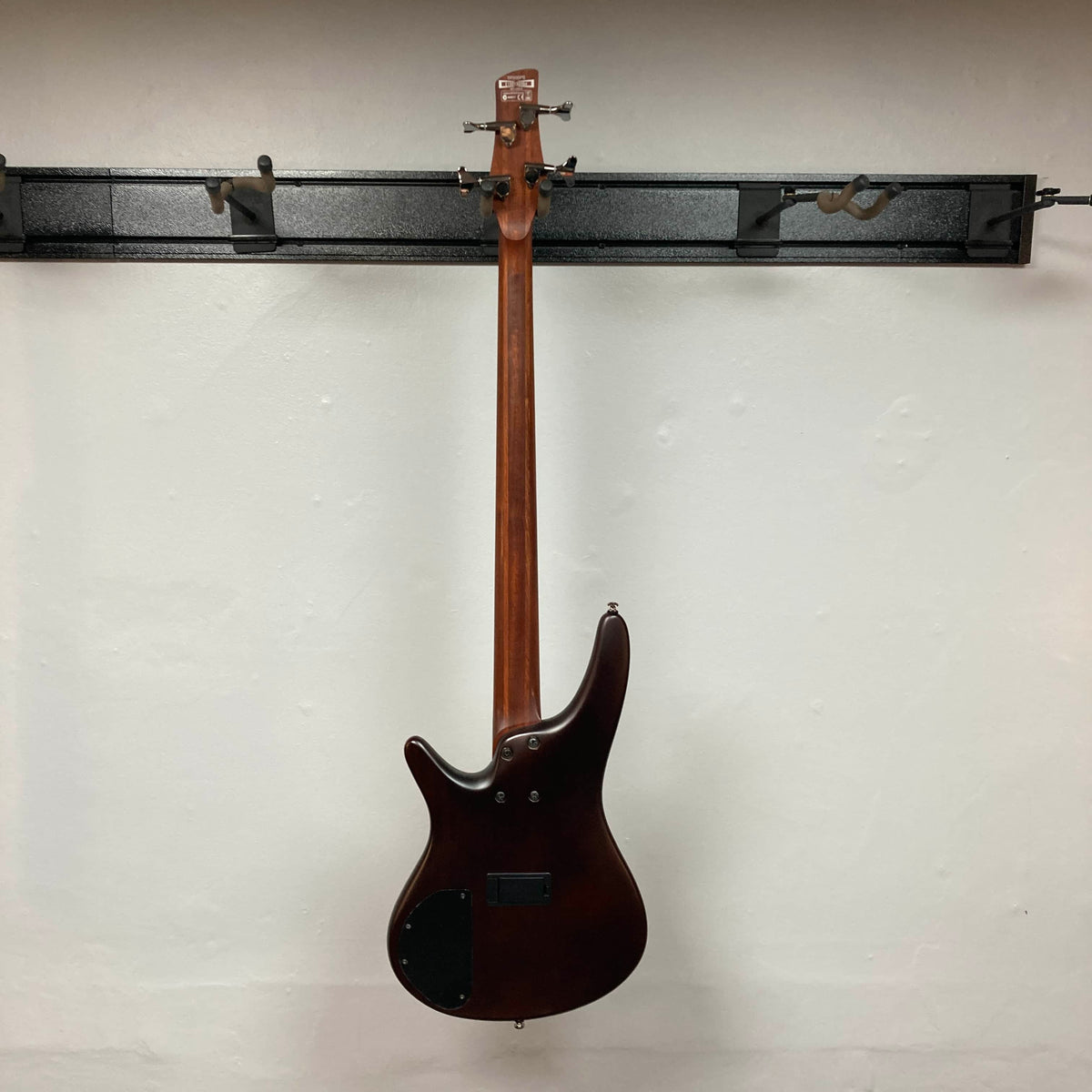 An Ibanez SDGR SR500PB bass guitar with hard case hangs on a wall-mounted rack, showing its back against a white wall.