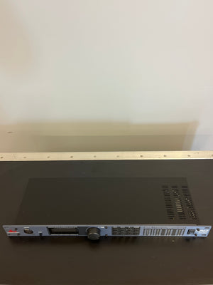 A dbx DriveRack PA Complete Loudspeaker Management System in black, featuring ventilation slots, buttons, and knobs, rests on a dark surface against a plain light-colored wall.