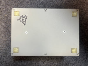 The Switchazel Boutique AB/Y Switcher with Tuner Out (Preowned) by Switchazel features a rectangular metal box, four rubber feet, and handwritten text on the bottom—perfect for guitar amp switching setups and eliminating ground loops.
