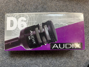 A box of the preowned Audix D6 Large Diaphragm Dynamic Kick Drum Microphone is displayed on gray carpet. The packaging features an image of the mic, designed for kick drums, and the Audix logo.
