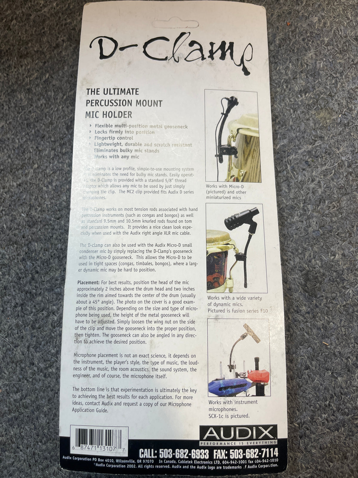 Back of an Audix D-Clamp Percussion Mount Mic Holder package by Audix, showing features, mounting instructions, compatibility details, and contact info on a gray carpet background.