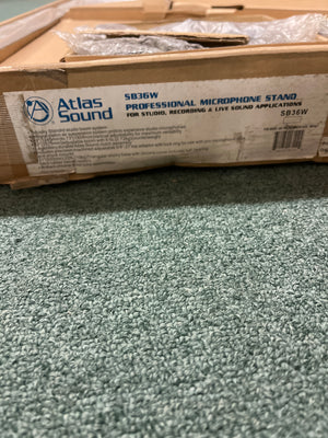 A cardboard box labeled "Atlas Sound SB36W Professional Studio Boom Microphone Stand" by Atlas Sound, featuring an air suspension system, sits on a textured green carpet.