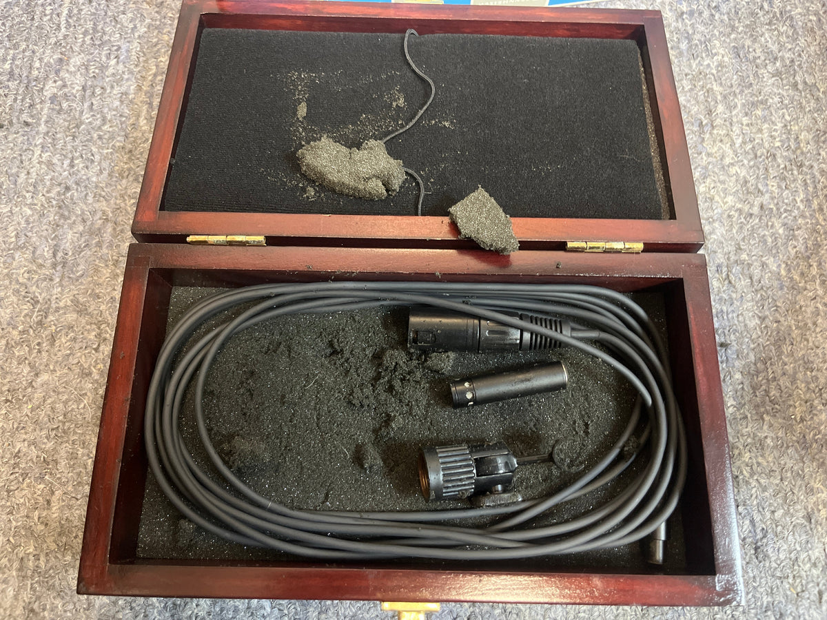 A wooden box with worn black foam holds a coiled audio cable and connectors, likely for the preowned Audix M1244-HC Miniature Condenser Hi-SPL Hypercardioid Microphone, resting on a textured gray surface.