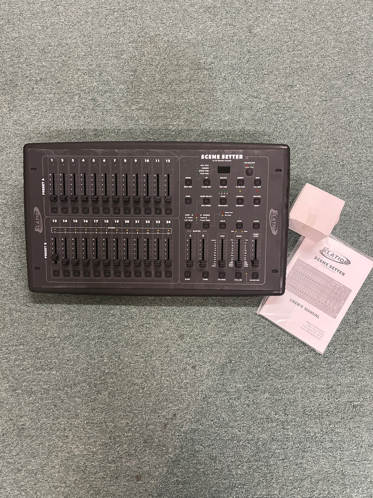 An Elation Scene Setter 24-Channel DMX Dimmer Console by Elation rests on a gray carpet, accompanied by its user manual and warranty card.