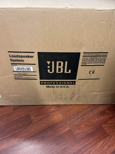 A cardboard box labeled "JBL Professional Loudspeaker System" sits on a wood floor, featuring the high-output VRX918S model for live sound reinforcement, with "Made in U.S.A." printed on the box.