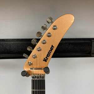 A close-up of a Kramer 84 HH Killswitch guitar headstock in Smoked Almond Metallic, featuring six tuning pegs, a black Kramer logo, and mounted on a wall holder against a plain light gray background. Includes gigbag.