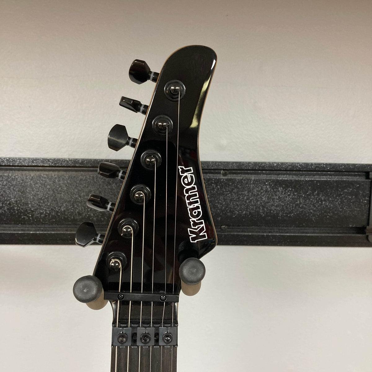Close-up of a black Kramer Volante Quilt HSFR headstock with six tuning pegs, Floyd Rose locking nut, and white Kramer logo, set against a light wall with a black horizontal rack in the background. Brand: Kramer. Color: Aqua Burst. Includes gigbag.