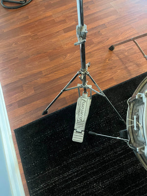 A hi-hat cymbal stand with a Ludwig foot pedal rests on a dark rug atop a wooden floor, next to the Ludwig Classic 1980s Drum Kit in White Marine Pearl.