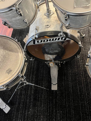 A close-up of a Ludwig Classic 1980s Drum Kit – White Marine Pearl shows the bass drum with Ludwig logo, drum pedal, and tom-toms on a black textured carpet.