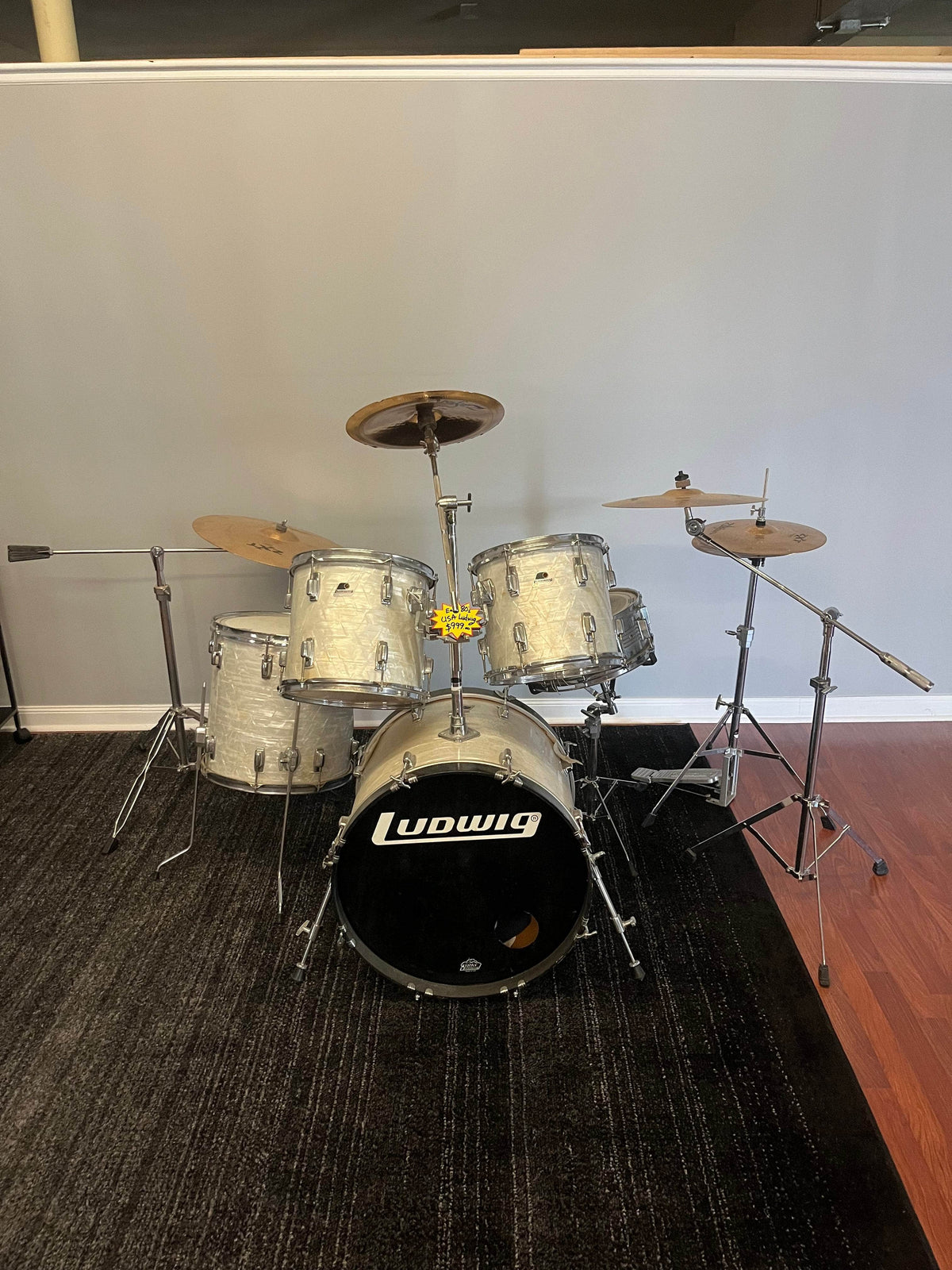 A vintage Ludwig Classic 1980s Drum Kit in White Marine Pearl is set on a dark carpet before a plain gray partition, cymbals included. A yellow flower rests atop the bass drum, adding charm to this classic Ludwig set.