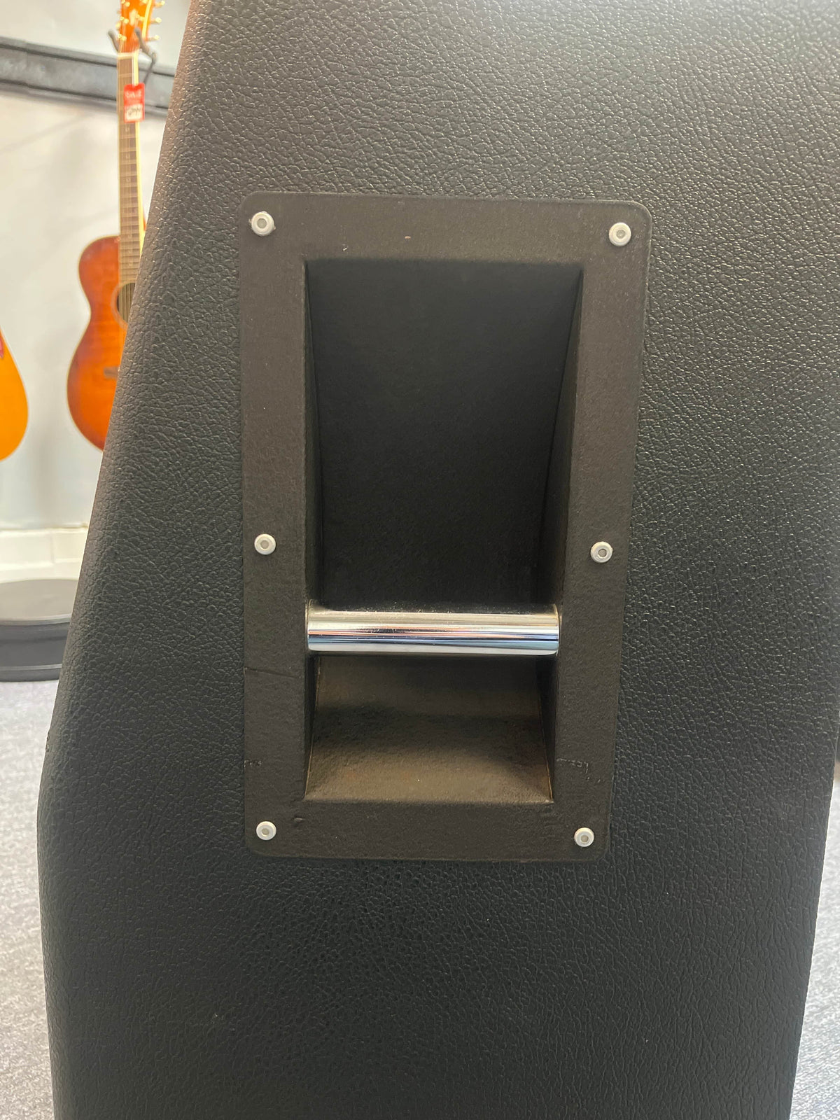 Close-up of the built-in metal handle on a black 1969 Marshall 4x12 Cabinet with Matching Celestion T1281 Greenbacks; blurred guitars appear in the background.