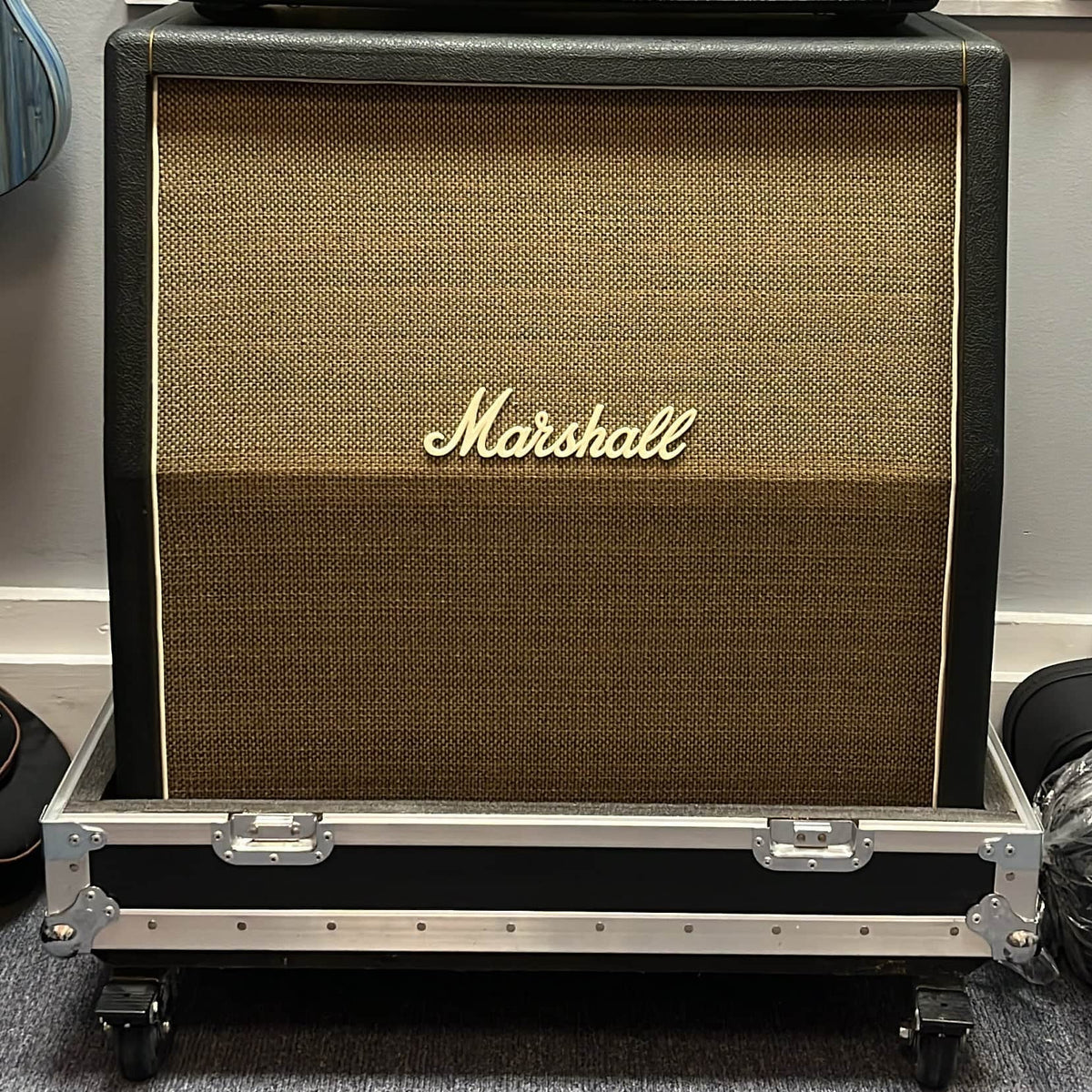 A 1969 Marshall 4x12 Cabinet with matching Celestion T1281 Greenbacks rests on a rolling flight case against a gray wall.