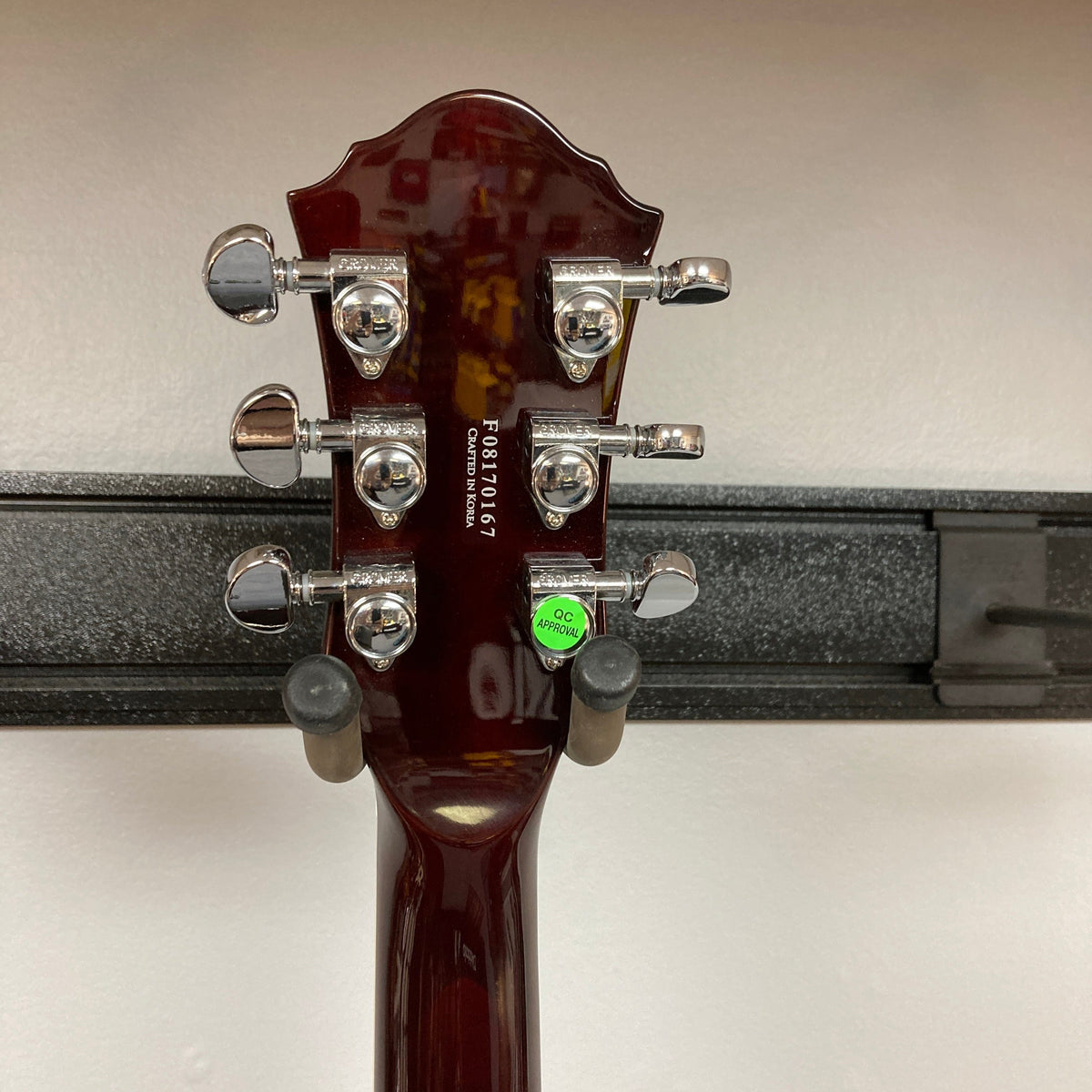 The back of a Michael Kelly Hybrid Special Electric Guitar - Spalt Burst headstock features six chrome tuning pegs, a serial number, and a green QC sticker. The guitar is on a black wall-mounted holder against a white background.