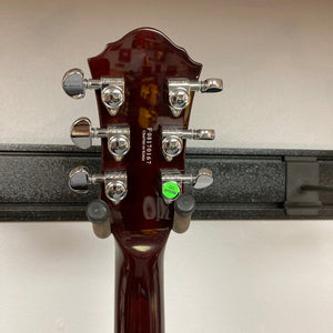 The back of a Michael Kelly Hybrid Special Electric Guitar - Spalt Burst headstock features six chrome tuning pegs, a serial number, and a green QC sticker. The guitar is on a black wall-mounted holder against a white background.