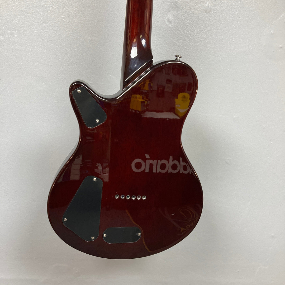 A close-up of the back of a Michael Kelly Hybrid Special Electric Guitar - Spalt Burst shows three black covers and six string holes on its glossy red finish, reflecting text and objects, set against a plain white wall.