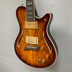 The Michael Kelly Hybrid Special Electric Guitar in Spalt Burst features a semi-hollow body, sunburst finish, dual humbuckers, two knobs, and an f-hole design—shown against a plain gray wall.