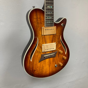 A Michael Kelly Hybrid Special Electric Guitar - Spalt Burst, featuring dual humbuckers, three wooden knobs, and f-hole cutouts in a glossy finish, hangs against a light gray wall.