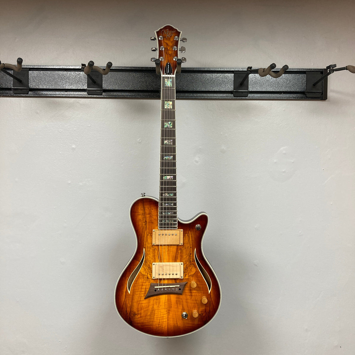 A Michael Kelly Hybrid Special Electric Guitar - Spalt Burst hangs on a black wall-mounted rack against a plain white wall. The guitar features two humbucker pickups and pearl fretboard inlays.