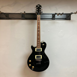 Michael Kelly Patriot Decree Lefty Black Blem electric guitar with gold knobs and chrome hardware displayed on a wall.