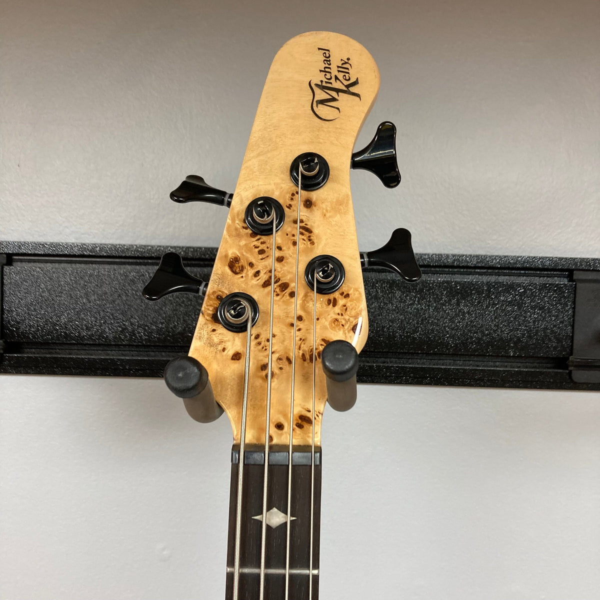 Michael Kelly Pinnacle 4 Bass neck and headstock on a black stand, showcasing tuning pegs and ornate design details.