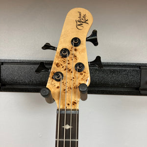 Michael Kelly Pinnacle 4 Bass neck and headstock on a black stand, showcasing tuning pegs and ornate design details.