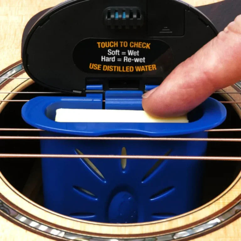 A person presses a yellow pad inside the blue Music Nomad The Humitar ONE Acoustic Guitar Humidifier & Hygrometer, which sits in an acoustic guitar’s sound hole. The lid has instructions for checking the pad's moisture level.