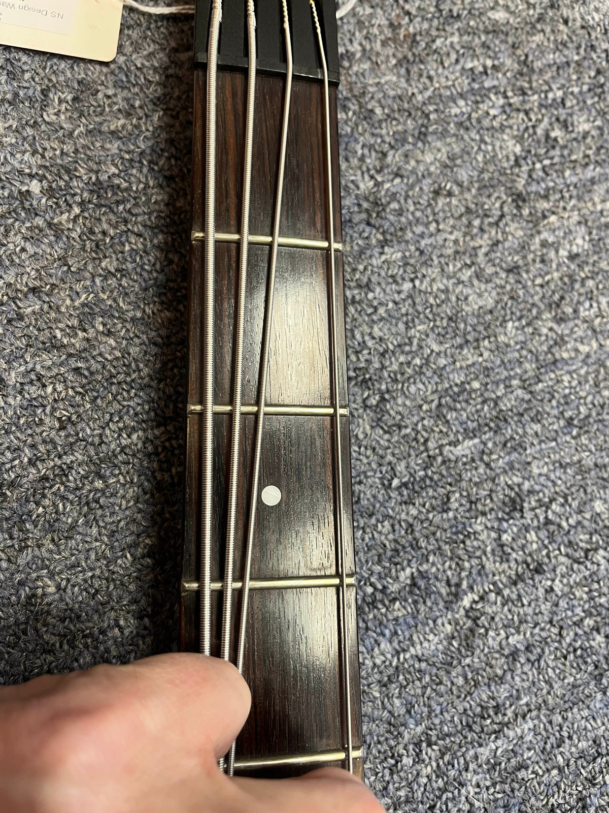 A close-up of a hand pressing on the strings of an NS Design Wav Radius Bass w/NS Gigbag, showcasing its punchy bass tones over a gray carpet.