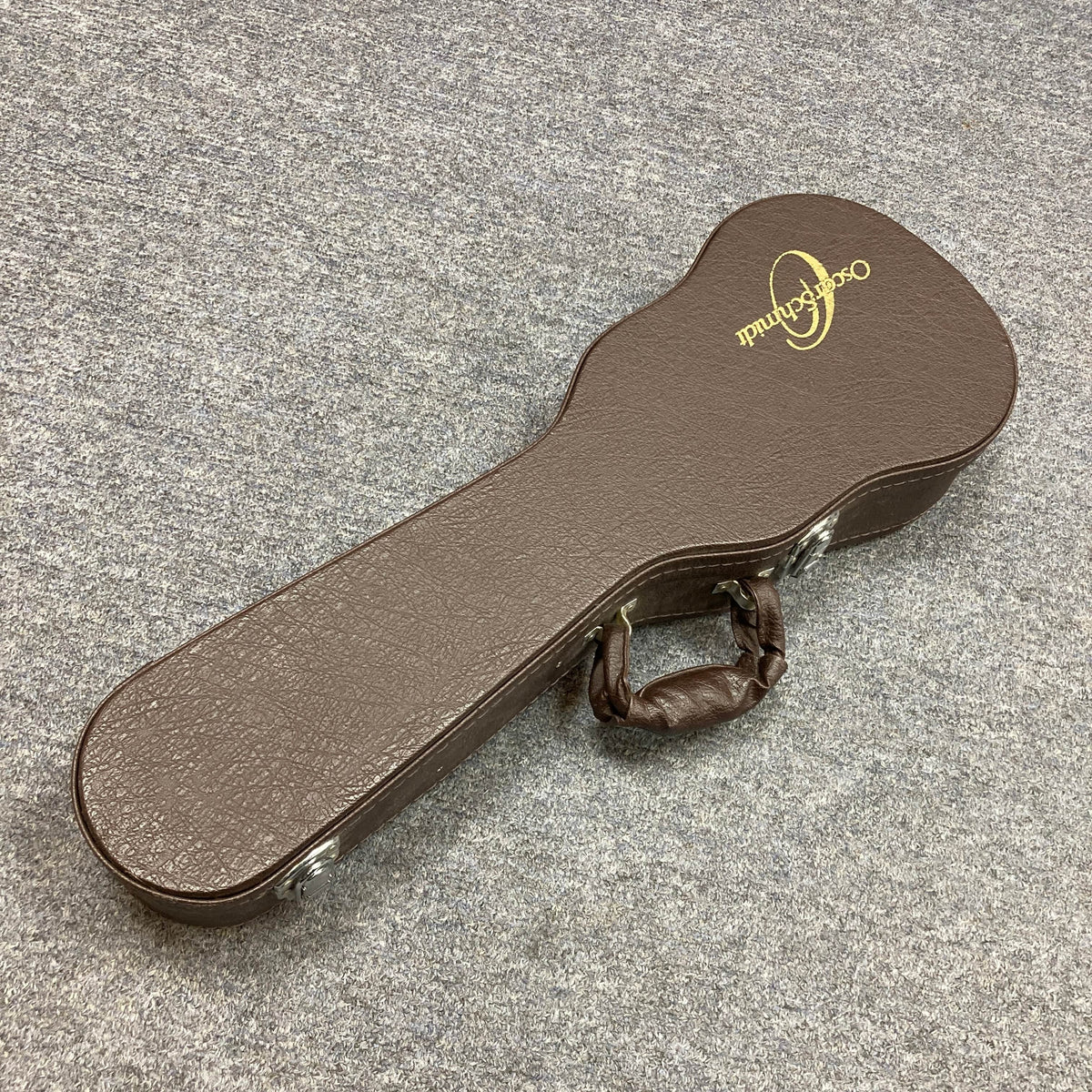 A brown hard-shell case with textured surface and gold Oscar Schmidt logo sits closed on a gray carpet, perfect for storing your Oscar Schmidt OU280SWK Concert Ukulele. The handle is wrapped in a matching brown cover.