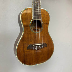 The Oscar Schmidt OU280SWK Concert Ukulele, a brown solid koa model with four strings and abalone binding, features a glossy finish and hangs on a light gray wall.