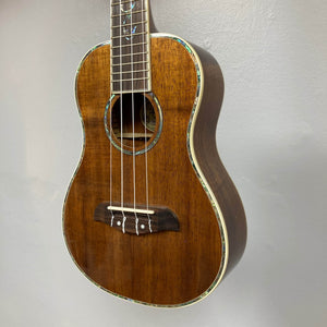 A close-up of an Oscar Schmidt OU280SWK Concert Ukulele with solid koa body, four strings, white fret markers, and abalone binding around the body and sound hole, hanging against a plain light gray wall. Includes case; used consign.