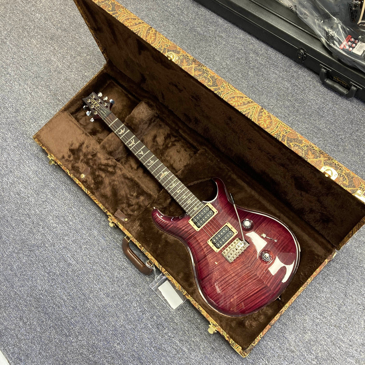A PRS Custom 24 Angry Larry 2011 electric guitar, featuring a striking flamed maple top in red, rests inside its open brown velvet-lined hard case on a carpeted floor, its gleaming PRS humbuckers catching the light.