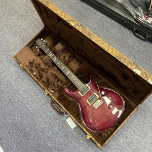 A PRS Custom 24 Angry Larry 2011 electric guitar, featuring a striking flamed maple top in red, rests inside its open brown velvet-lined hard case on a carpeted floor, its gleaming PRS humbuckers catching the light.