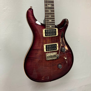 A PRS Custom 24 Angry Larry 2011 electric guitar by PRS, featuring bird inlays on the fretboard, two humbucker pickups, a tremolo bridge, and control knobs, is hanging on a light gray wall.
