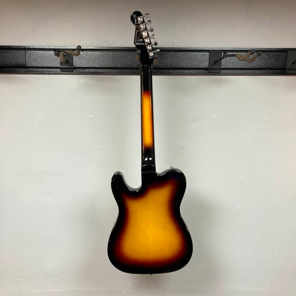 A Reverend Greg Koch Signature Gristle 90 electric guitar in 3-Tone Burst is hanging on a wall rack, photographed from the back against a plain white wall.