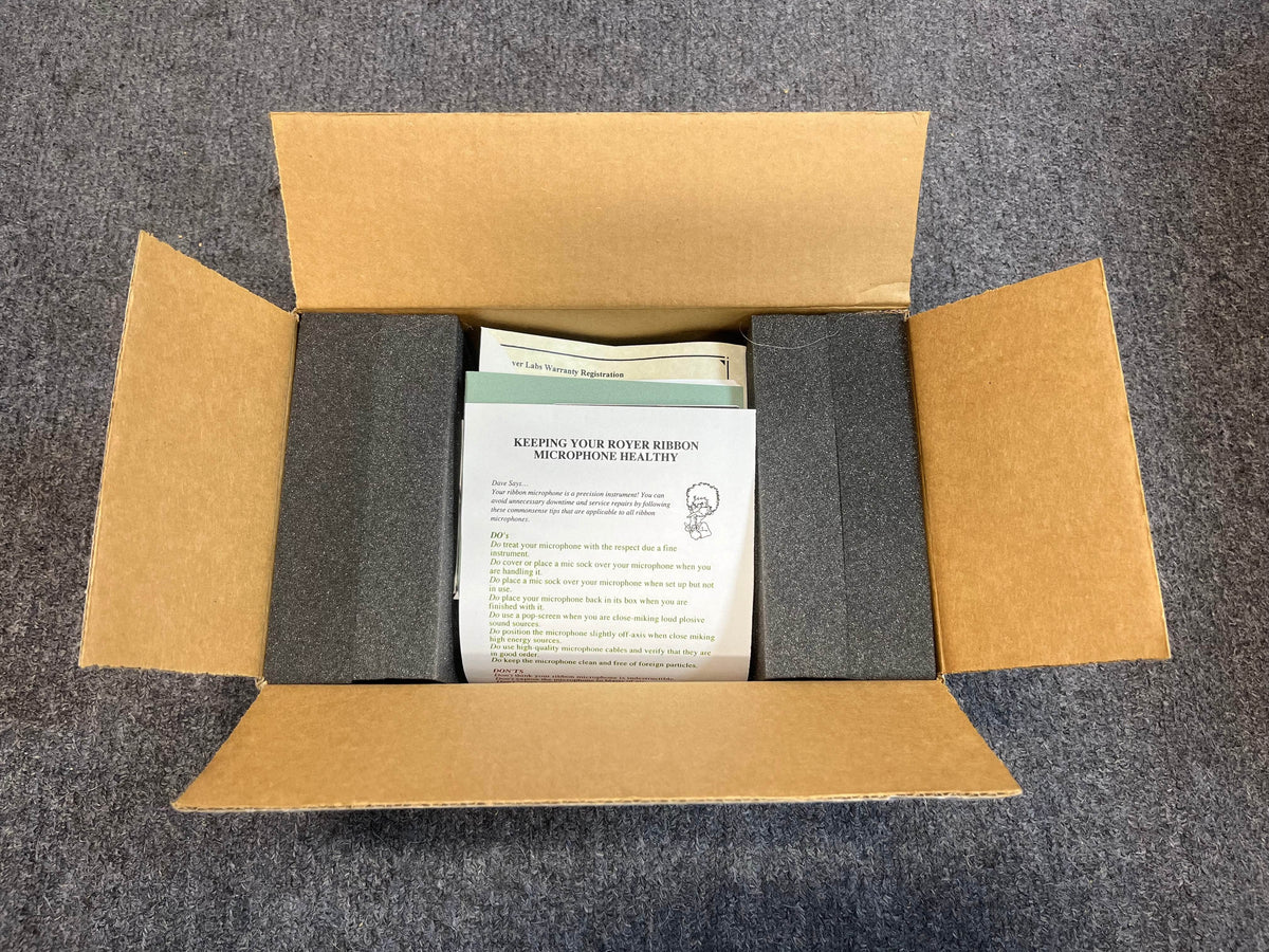 An open cardboard box on a gray carpet holds documents and foam padding, with an instruction sheet on top—ideal for anyone using the Royer Labs R-121 Ribbon Microphone (Pre-owned in excellent condition) by Royer Labs.