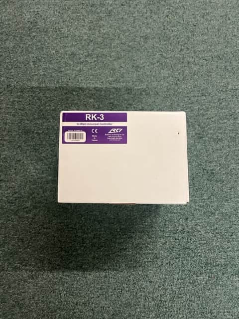 A white rectangular RTI RK-3 In-Wall Universal Controller by Remote Technologies Inc sits on green carpet, perfect as a home automation in-wall controller.