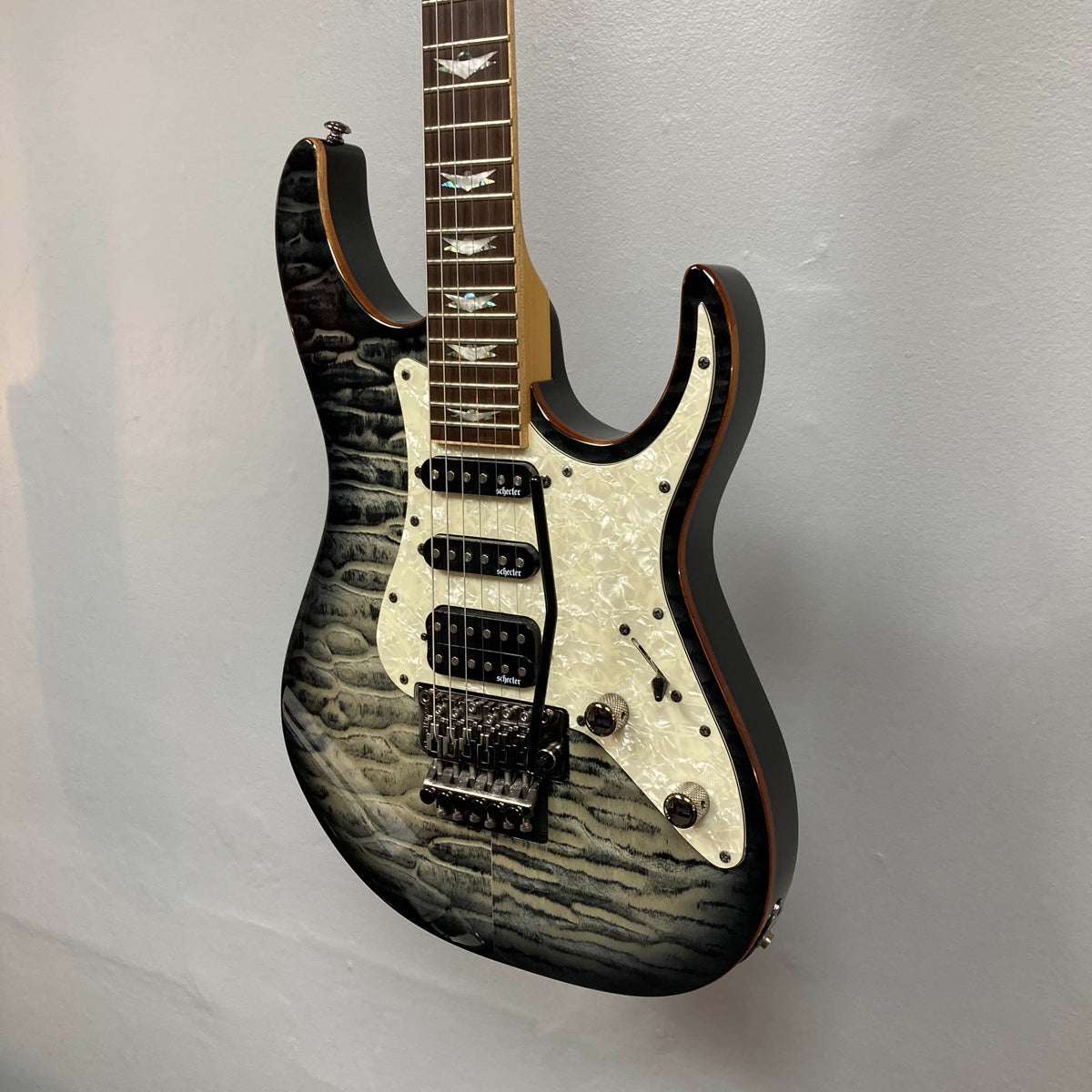 The Schecter Extreme Banshee 6 FR CHARCOAL BURST electric guitar with case by Schecter is shown mounted on a light-colored wall, featuring high-output electronics, three pickups, and a Floyd Rose tremolo bridge.