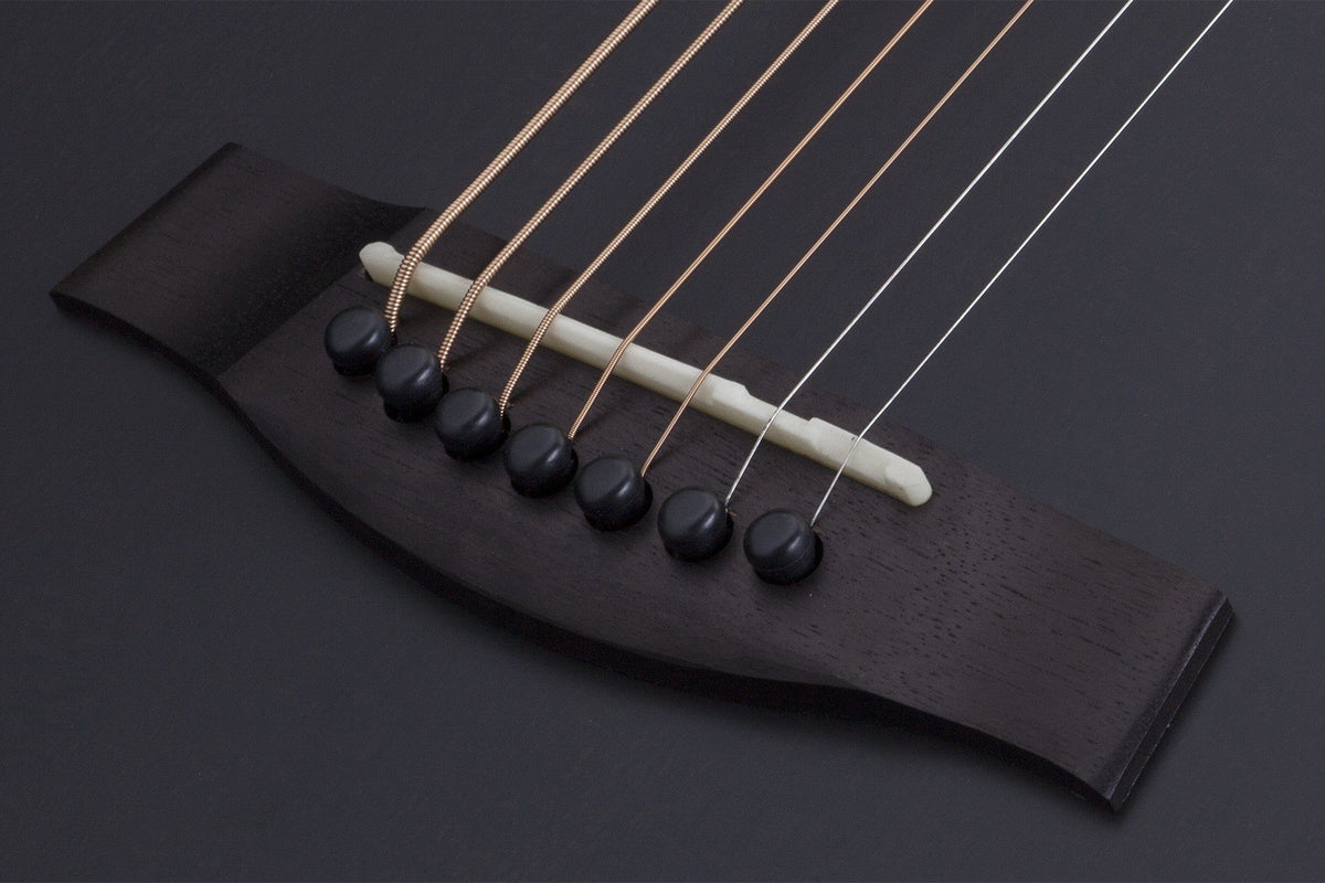 Close-up of the Schecter Orleans Stage 7-String Acoustic Guitar bridge, featuring six black pins, six strings, and a white saddle on dark wood against a black background—ideal for extended range by Schecter.