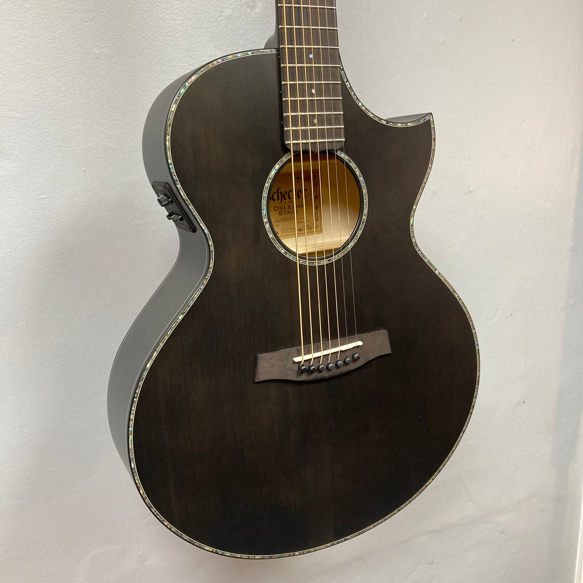A Schecter Orleans Stage 7-String Acoustic Guitar in black with a cutaway body, abalone binding, Engelmann spruce top, and visible sound hole hangs against a plain light-colored wall.