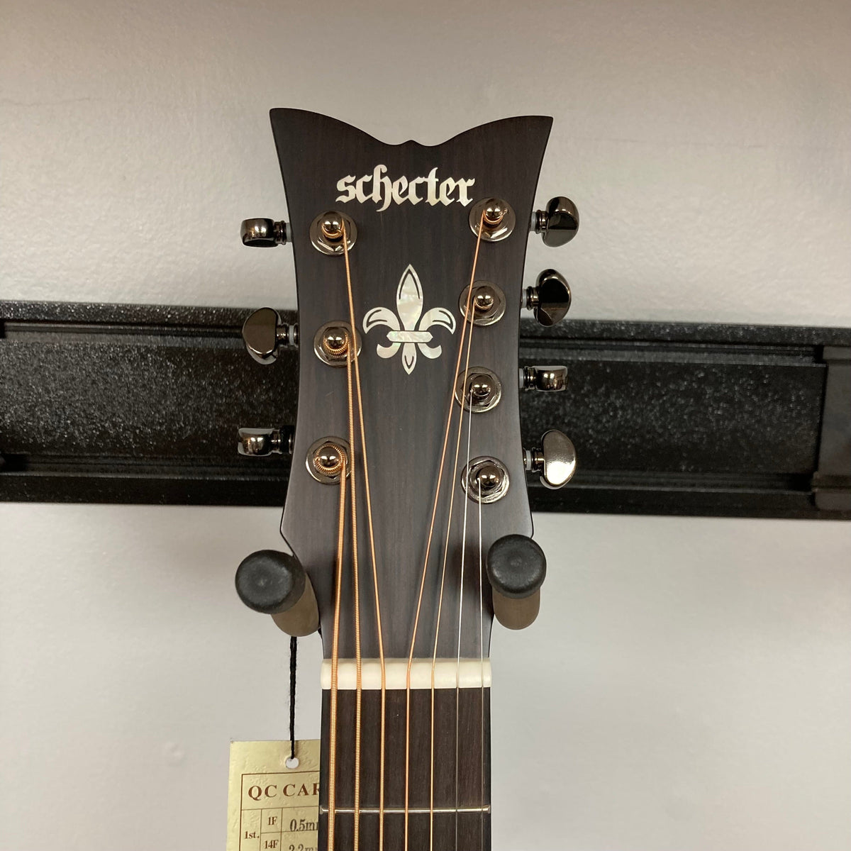 The headstock of a Schecter Orleans Stage 7-String Acoustic Guitar features seven tuning pegs, a fleur-de-lis inlay, and a tag on one string; it’s shown on a wall hanger against a light background.