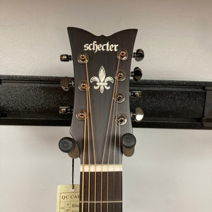 The headstock of a Schecter Orleans Stage 7-String Acoustic Guitar features seven tuning pegs, a fleur-de-lis inlay, and a tag on one string; it’s shown on a wall hanger against a light background.