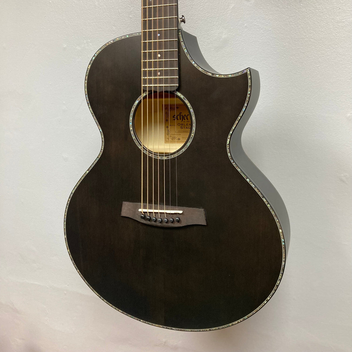 The Schecter Orleans Stage 7-String Acoustic Guitar, shown in black with a cutaway body against a light gray wall, features decorative binding, modern style, and the extended range of seven strings.