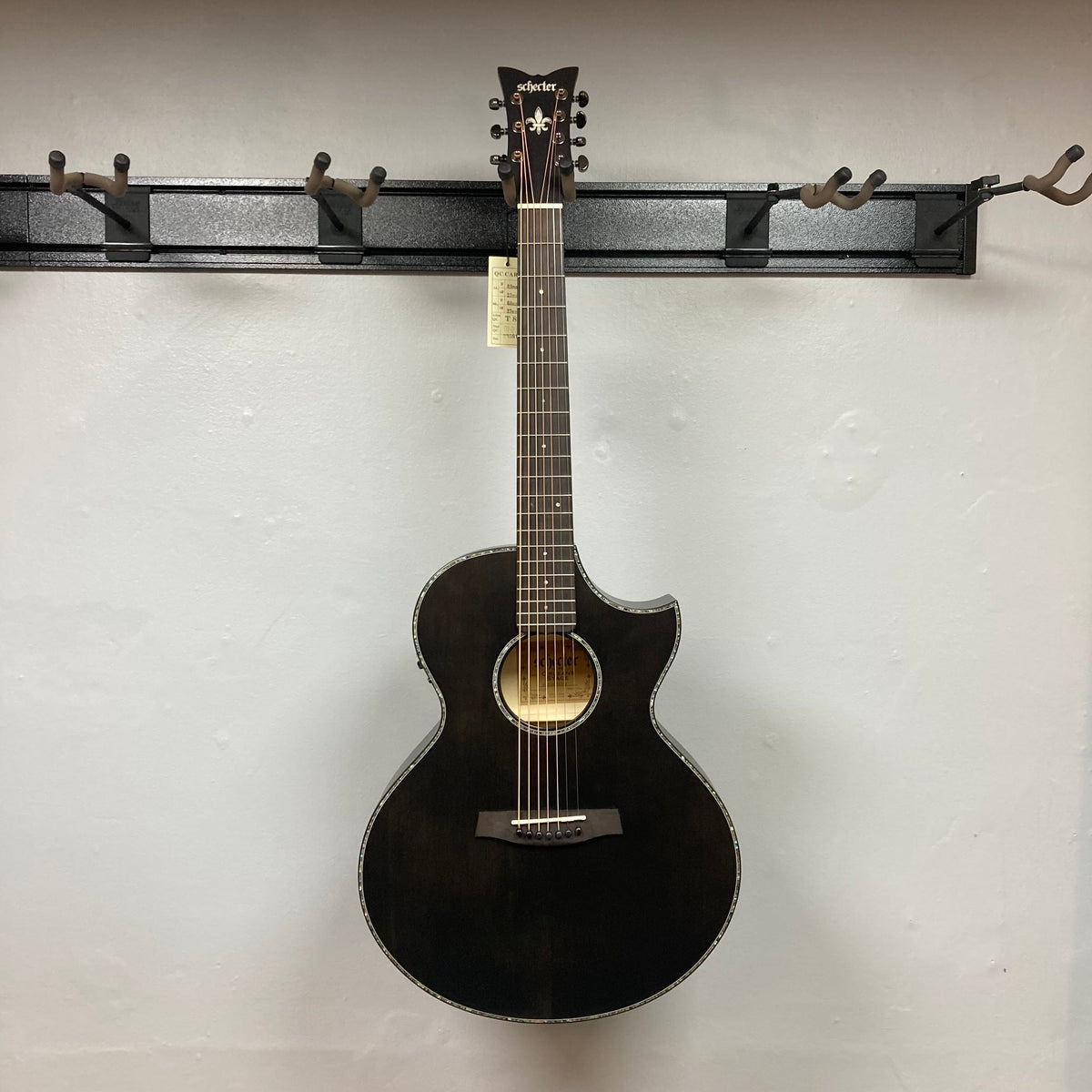 A Schecter Orleans Stage 7-String Acoustic Guitar in black, featuring a cutaway body and Engelmann spruce top, hangs on a wall-mounted rack against a plain white background. The headstock shows the Schecter brand label.