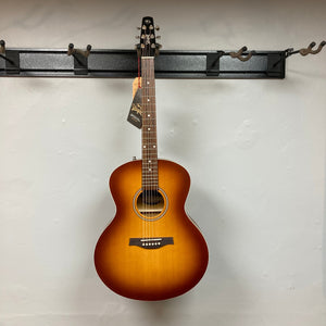 Seagull Entourage Mini Jumbo Rustic Burst MJ guitar hanging on a wall, featuring a tag on the neck and detailed wood grain finish.
