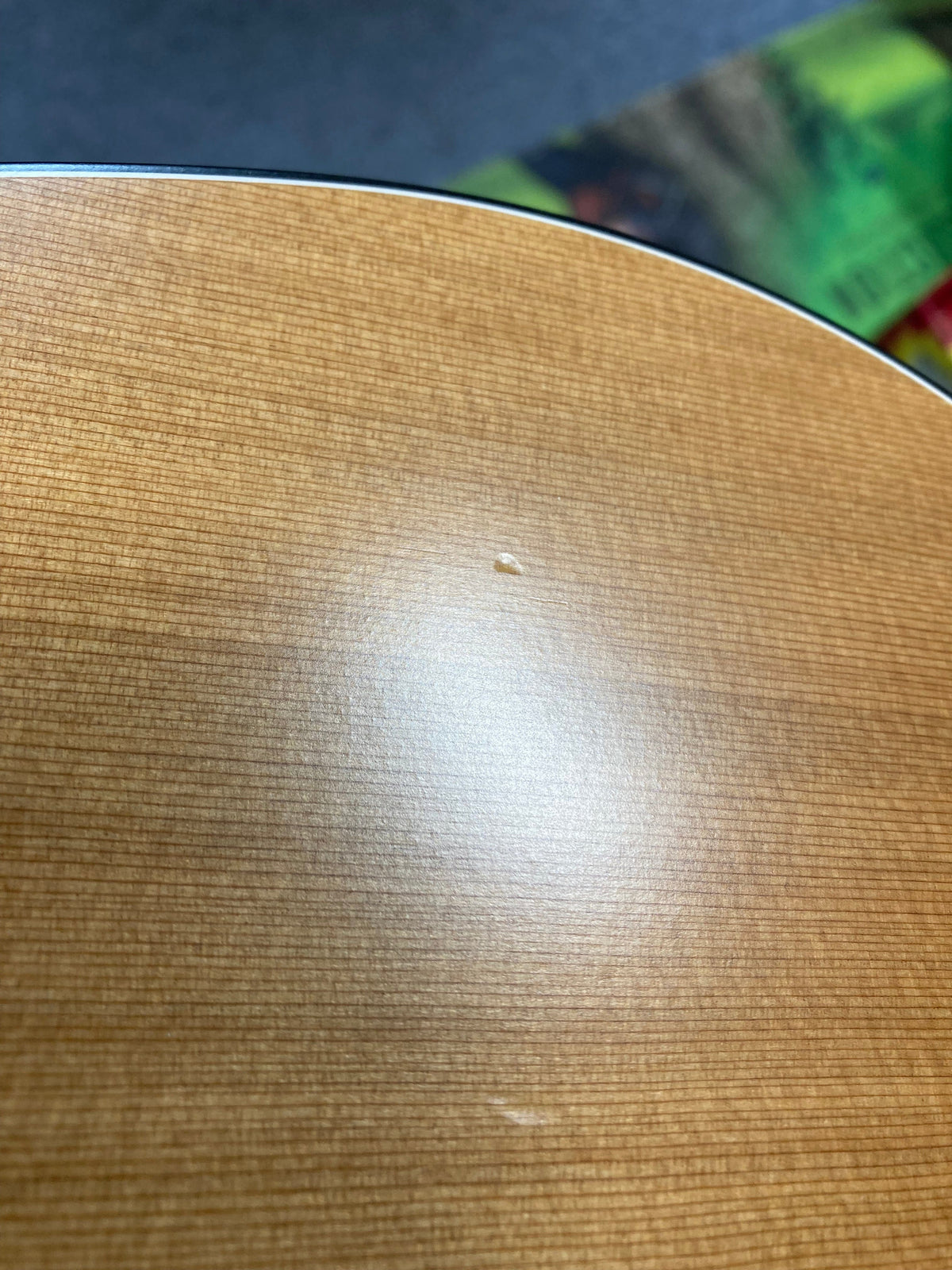 Close-up of a Seagull S6 Original Left Handed Acoustic Guitar (Blemished) cedar top by Seagull, showing light brown wood with a small dent and tiny blemish near the center; blurred green and red object in background.