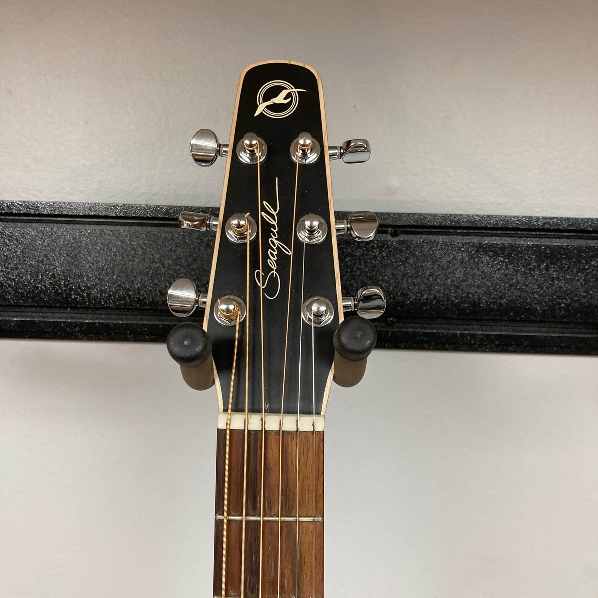 The headstock of a Seagull S6 QI Slim Acoustic Guitar w/Hard Case by Seagull, showing six tuning pegs, is mounted on a black wall hanger.