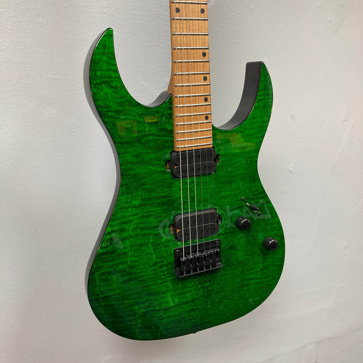 The Spira S-450 Trans Green TGR, a modern green electric rock and metal guitar by Spira with a roasted maple neck and black custom humbuckers, is mounted on a white wall. Its contoured body has two control knobs near the bridge.