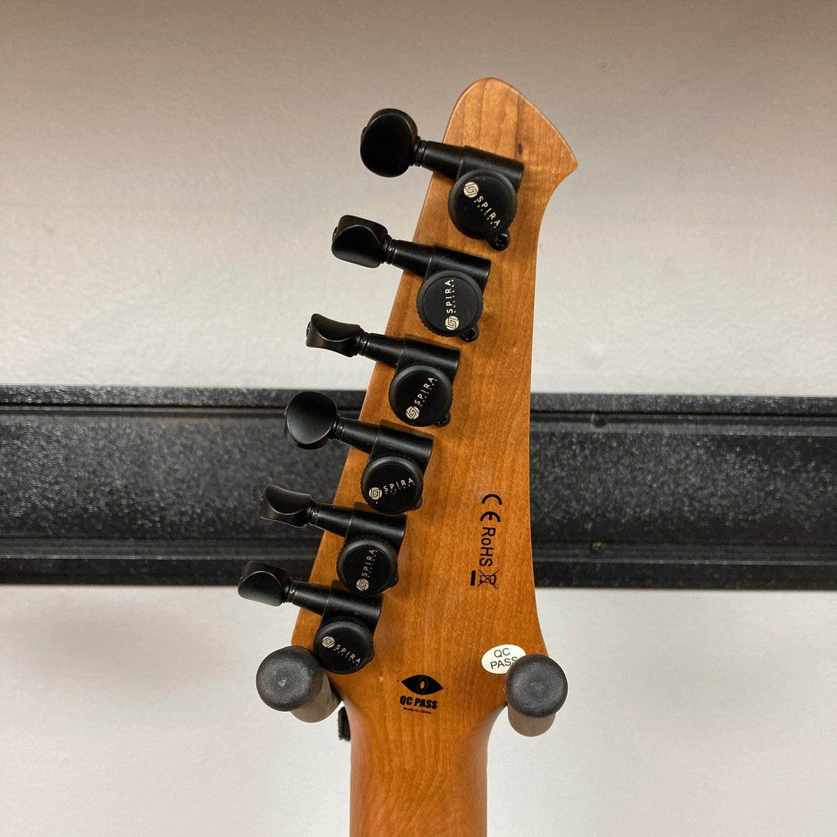 The back of a Spira S-450 Trans Green TGR's headstock features six black tuning pegs, manufacturer labels, and certification markings. Its roasted maple neck hints at quality, set against a black horizontal strip on a white wall.