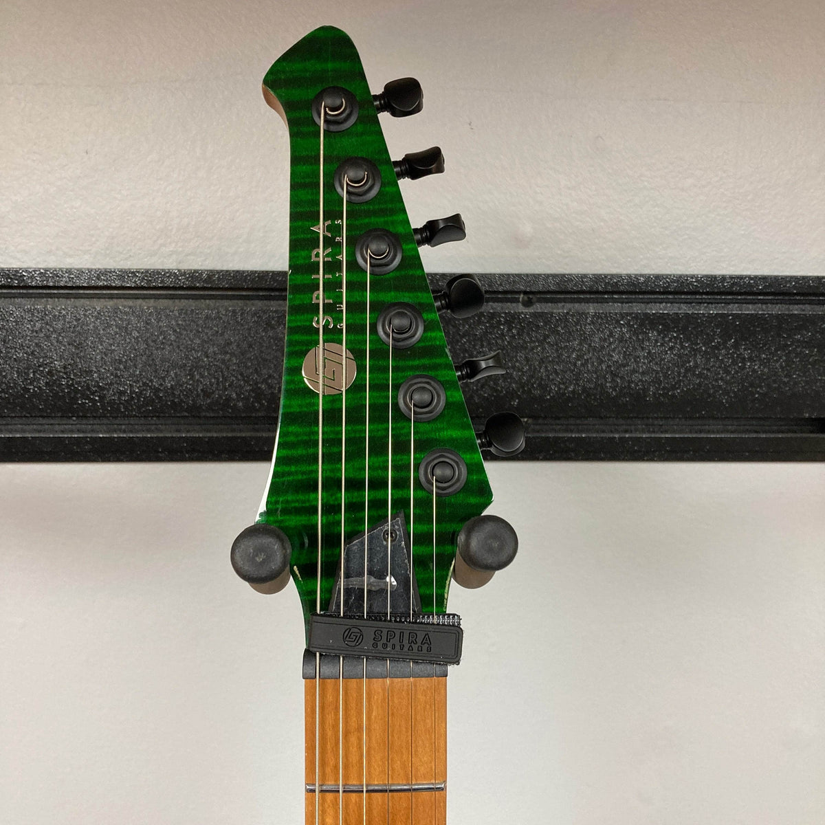 A close-up of the Spira S-450 Trans Green TGR headstock shows its green stripes, six tuning pegs, locking nut, and “Spira” branding. Its roasted maple neck stands out against a white wall and black mounting bracket.